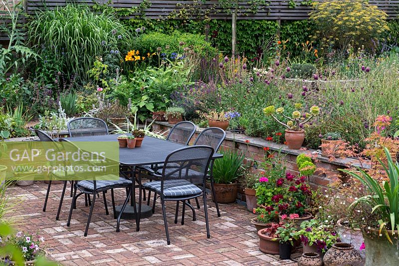 An outdoor dining table and chairs on the brick terrace, with retaining wall, mixed perennials and container planting.
 