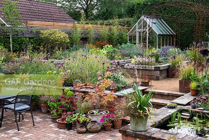 A split level, enclosed village garden with raised wooden beds planted largely with herbs, borders of late flowering summer perennials, a raised pond, a sunken terrace, pots of succulents and ornamental grasses, a wrought iron pergola and greenhouse.