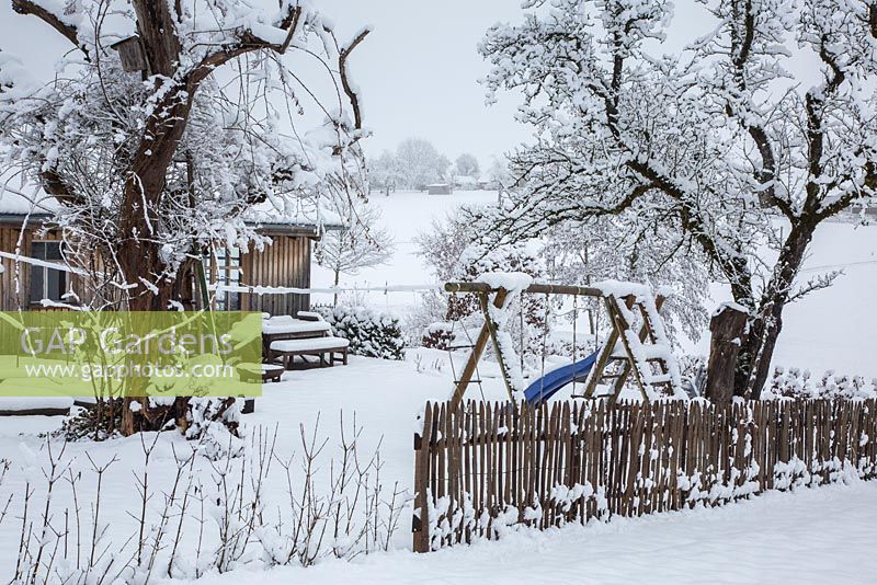 Winter scene with wooden picket fence, children's play equipment, trees and a shed