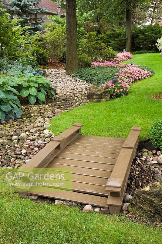 Manicured green grass lawn with brown painted wooden footbridge over dry stream and borders with Hosta plants and pink Impatiens flowers in residential backyard garden in summer