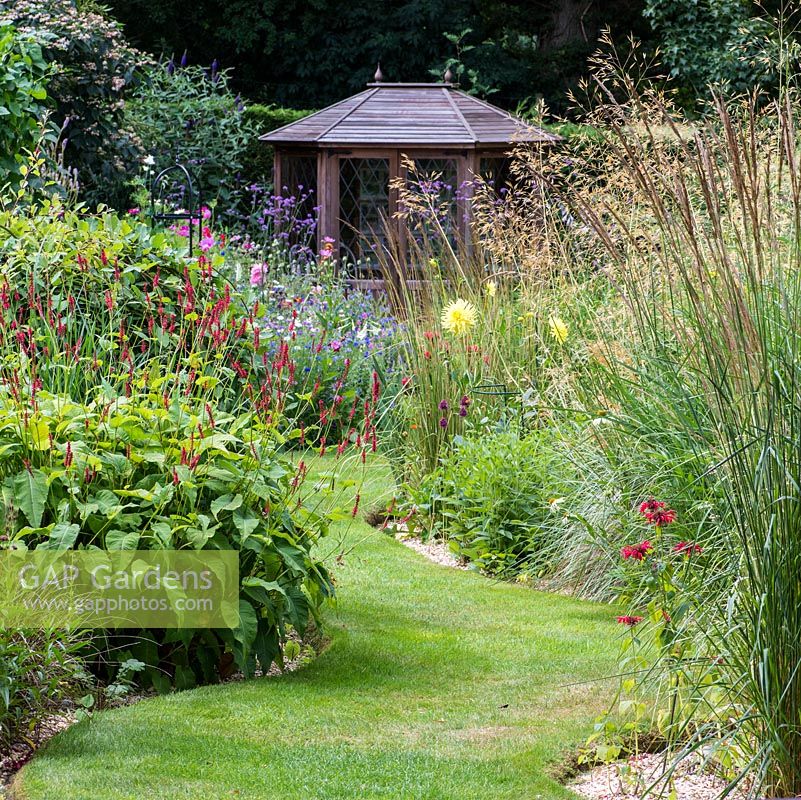 A grass path leads to a summerhouse  between herbaceous borders planted with Monarda, Persicaria, Dahlia, Verbena bonariensis with Stipa and Calamagrostis grasses.