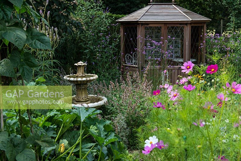 A stone water feature and wooden summerhouse amongst a border planted with Cosmos, Verbena bonariensis and Salvia.