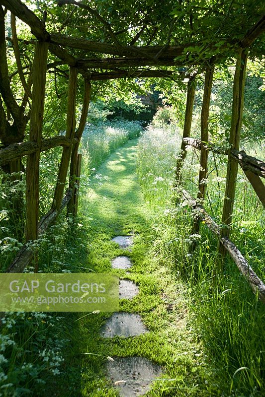Grassy mown paths lead beneath wooden arbour into the meadow and along a line of espaliered fruit trees toward the formal garden. King John's Nursery, Etchingham, East Sussex, UK