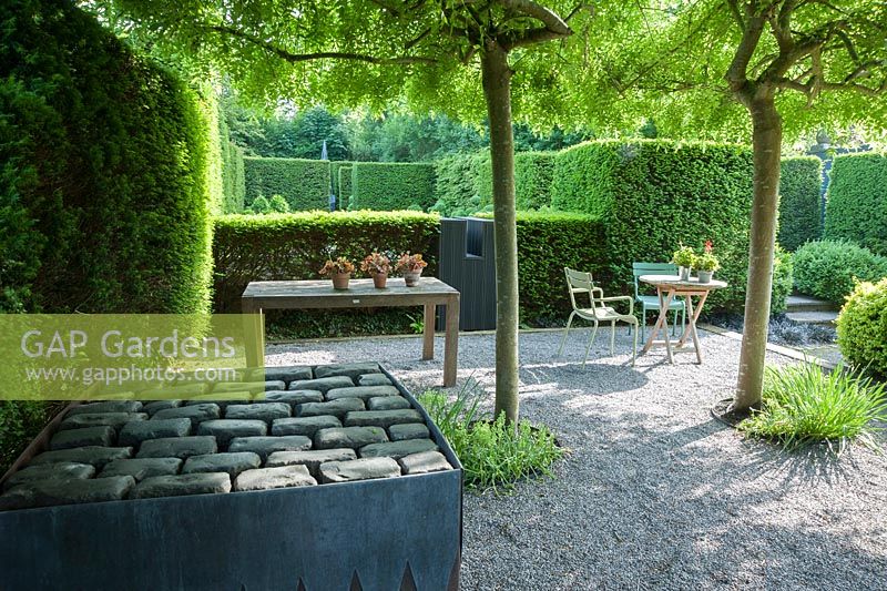 Graveled courtyard area beside the studio with four weeping ash, Fraxinus excelsior 'Pendula' forming a shady canopy, steel and lead container, filled with an arrangement of stone setts and a seating area surrounded by yew hedging.