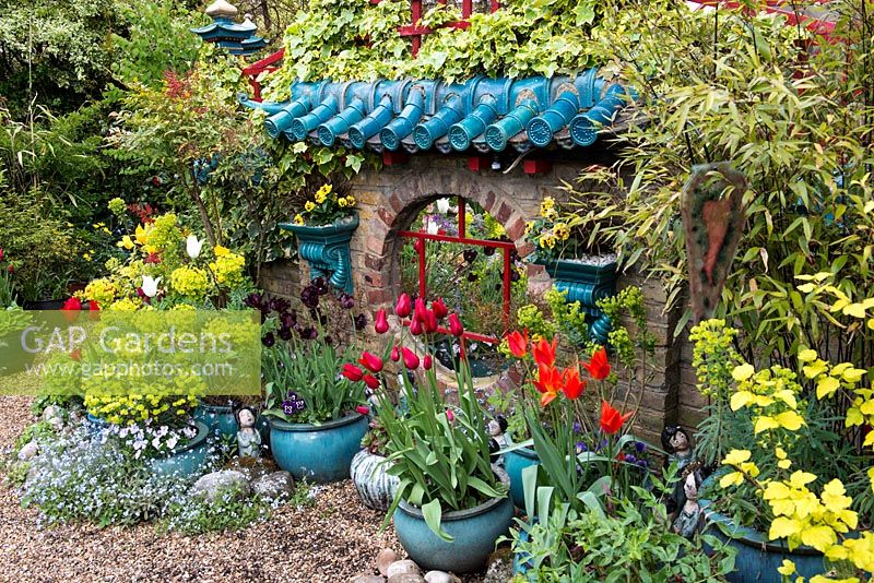 Oriental raku-tiled mirrored wall by artist owner, edged in bamboo, Nandina domestica and ivy. Surrounded by pots of tulips, self-seeded forget-me-not and euphorbia.
