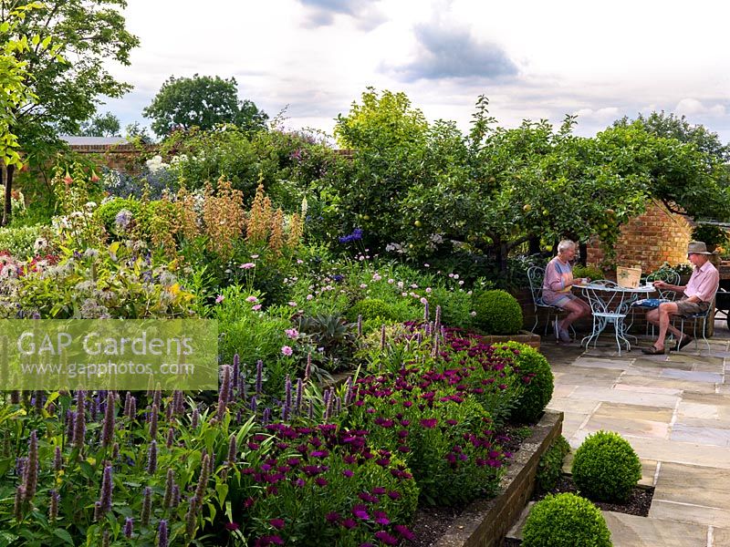 Owners Gayle and John Leader drink tea in the shade of mature apple tree, on sunken terrace edged in beds of osteospermum, agastache, clematis, agapanthus and box balls. Stuart Cottage.