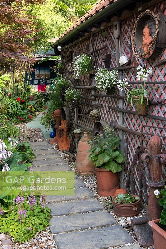 A town garden with narrow slab path besides a painted garage decorated with salvaged garden tools and hanging containers.