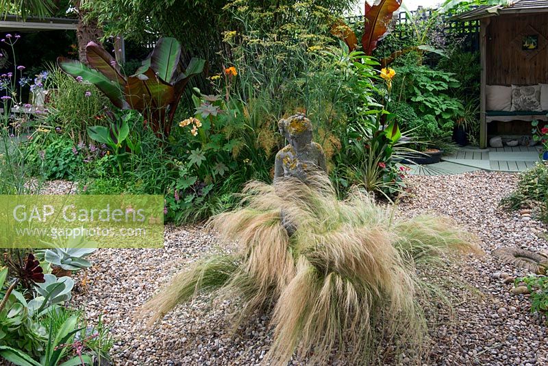 A gravel garden with Stipa grasses surrounding a stone statue in front a border with tropical foliage plants.