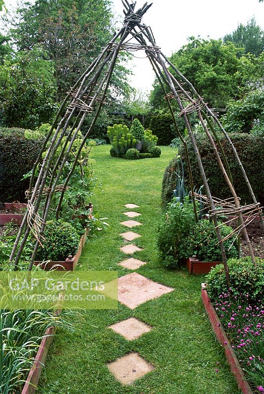 Rustic hazel arch frames garden. Path through wooden edged vegetable and herb beds with diamond shaped stepping stones 