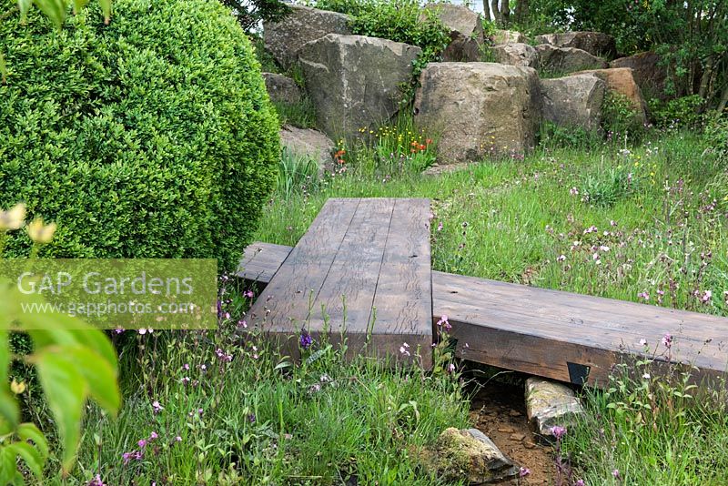Wooden boardwalk over stream, boulders, box ball and wildflowers. The Laurent-Perrier Chatsworth Garden. RHS Chelsea Flower Show 2015