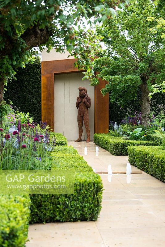 The Morgan Stanley Healthy Cities Garden. Sandstone path with water feature leading to steel sculpture of man with a child surrounded by symmetric hedges - Buxus sempervirens, fern - Dryopteris affinis, Salvia nemorosa 'Caradonna', Camassia cusickii and Lupinus 'Masterpiece'. 