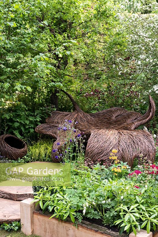 View of a woven willow seat in a tranquil woodland glade. Breast Cancer Haven Garden. RHS Chelsea Flower Show 2015