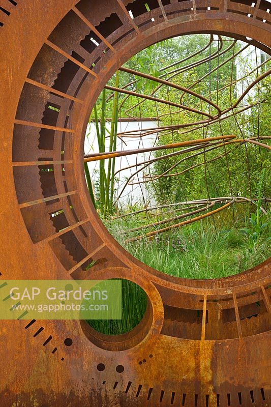 A rusted corten steel oculus and warped steel rods depicting the trajectory of light flow through the garden planted with foliage plants including Phyllostachys and grasses. Dark Matter Garden for the National Schools' Observatory. Design. RHS Chelsea Flower Show. 