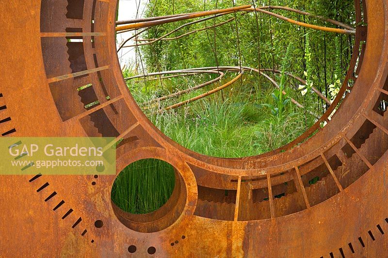 A rusted corten steel oculus and warped steel rods depicting the trajectory of light flow through the garden planted with foliage plants including Phyllostachys and grasses. Dark Matter Garden for the National Schools' Observatory. RHS Chelsea Flower Show 2015. 