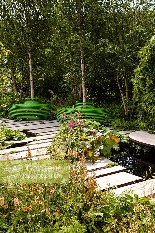 The Macmillan Legacy Garden - view showing tree seats, stepping stones across a pool and pond with edge of woodland planting including Rehmannia Walbertons 'Magic Dragon', Verbascum and Geum 'Totally Tangerine'  - RHS Hampton Court Flower Show 2015