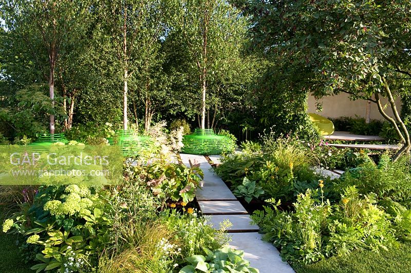  White stone paving through shady woodland-edge garden with pod structure seating area. Three birch trees with green-painted seats. The Macmillan Legacy Garden Sponsor Macmillan Cancer Support. RHS Hampton Court Palace Flower Show 2015