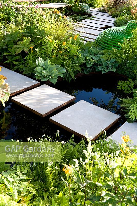 White slab stepping stones over black-water pool with naturalistic green, shade tolerant planting. The Macmillan Legacy Garden Sponsor Macmillan Cancer Support. RHS Hampton Court Palace Flower Show 2015