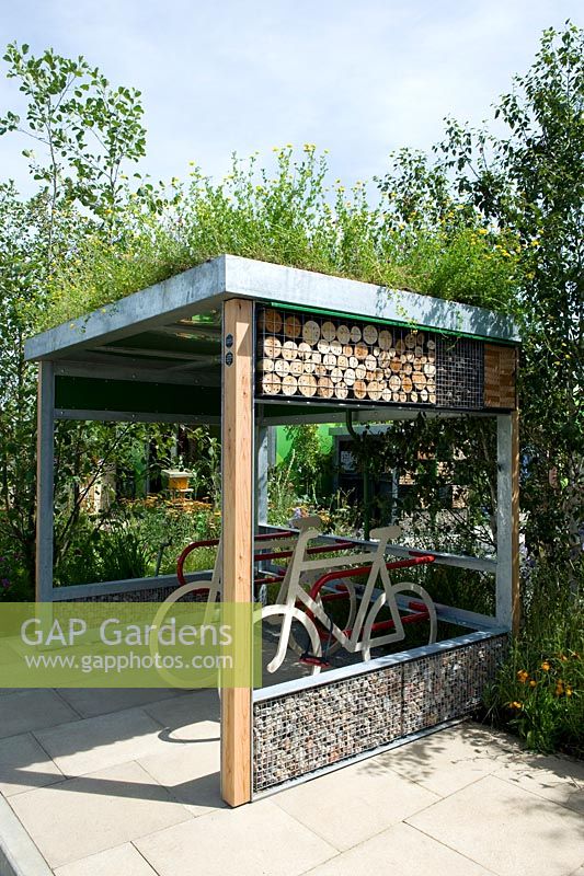 Urban street bicycle shelter with green roof backed by trees.  Community Street. Sponsor RHS, Gardeners' Question Time. RHS Hampton Court Flower Show, 2015.