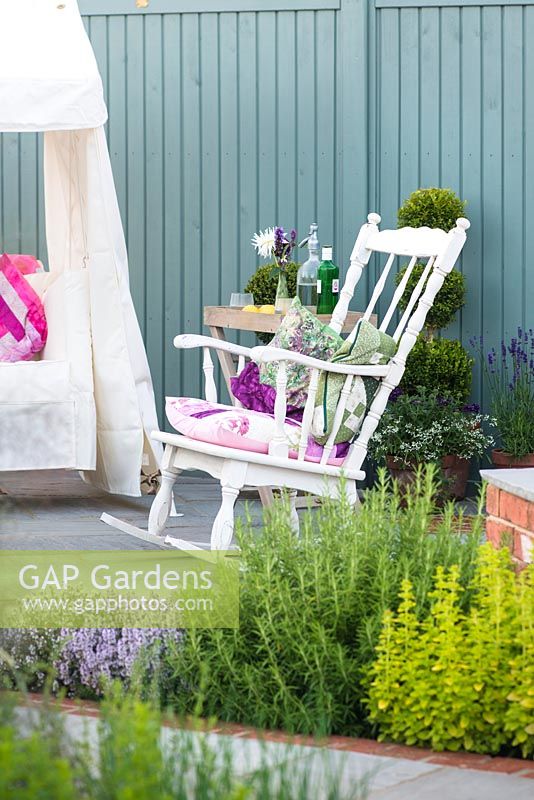 Just Retirement: A Garden For Every Retiree, view of seating area with white rocking chair against blue wooden wall surrounded by bed with herbs. Designer: Tracy Foster Sponsor:  Just Retirement Ltd