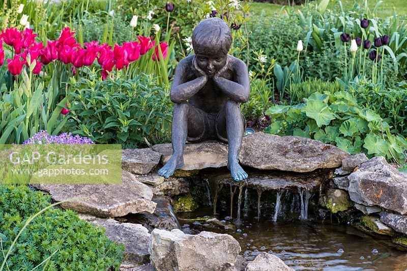 Figurine of a contemplative child, sculpted by Jenny Wynn Jones, overlooks a stone pool. Behind, red Tulipa 'Pimpernel'.