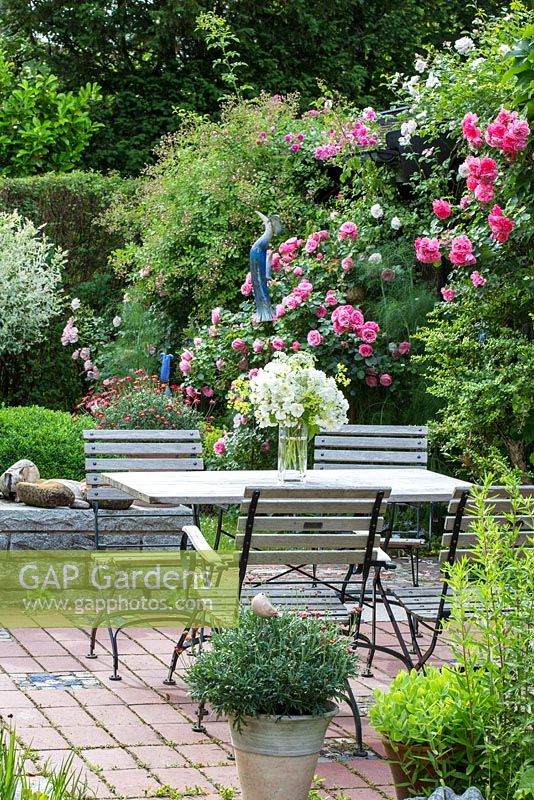 Wood and iron garden furniture on a brick stone terrace in a pottery maker's garden with ceramic objects and Rosa 'Leonardo da Vinci', 'New Dawn' and 'Rosarium Uetersen' 