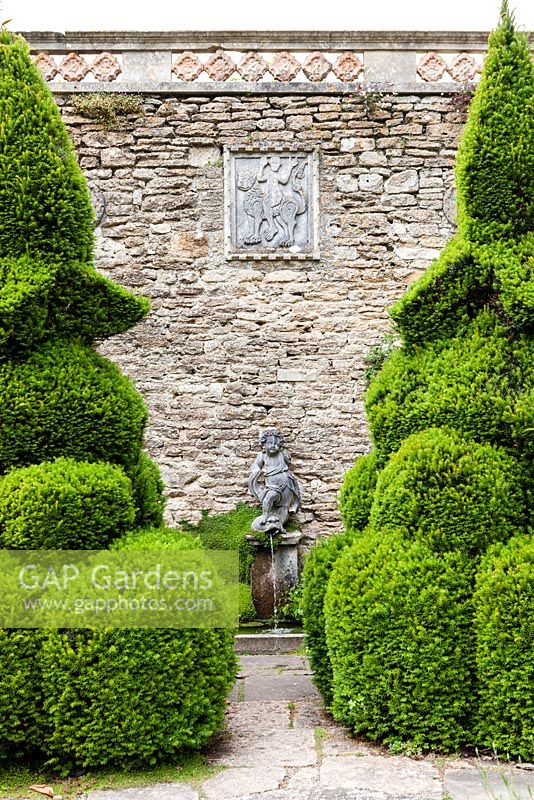 Clipped yews frame the Blue Pool, a small formal pool, with a Romanesque bas relief set into the wall above of a woman riding a lion. Iford manor, Bradford-on-Avon, Wiltshire. July. Garden designed and created by Harold Peto. Historic garden grade I.