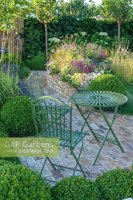 Wrought iron table and chairs on brick paving surrounded by box topiary balls - Squire's Garden Centres Urban Oasis - RHS Hampton Court Flower Show 2015