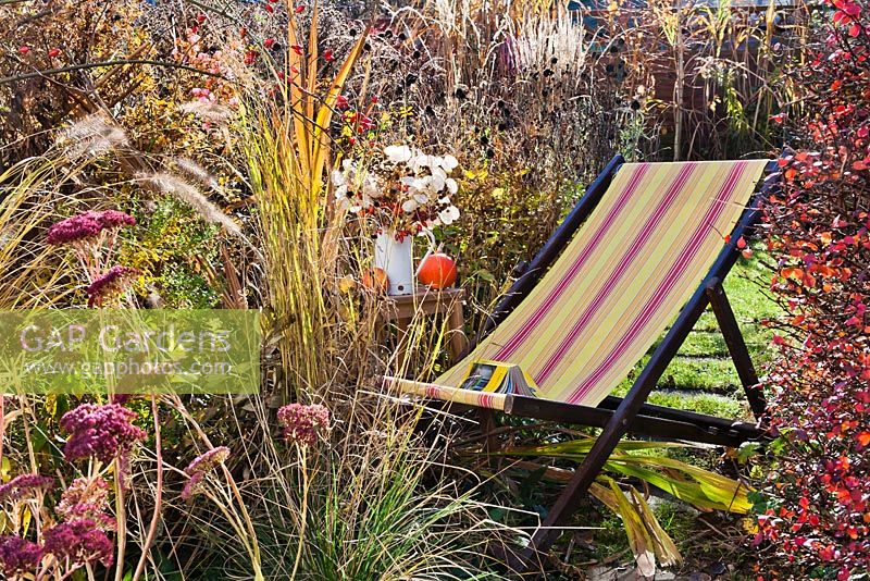 Deck chair amongs perennials and grasses in autumn. Pennisetum, Crocosmia 'Lucifer', Aster, Sedum, Panicum virgatum.