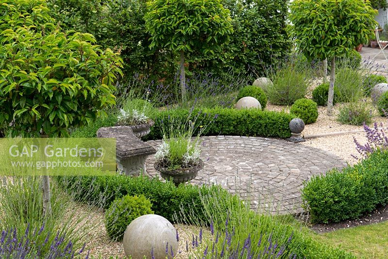 A circular patio with stone bench surrounded by low box hedging, portuguese laurel standards and stone spheres.