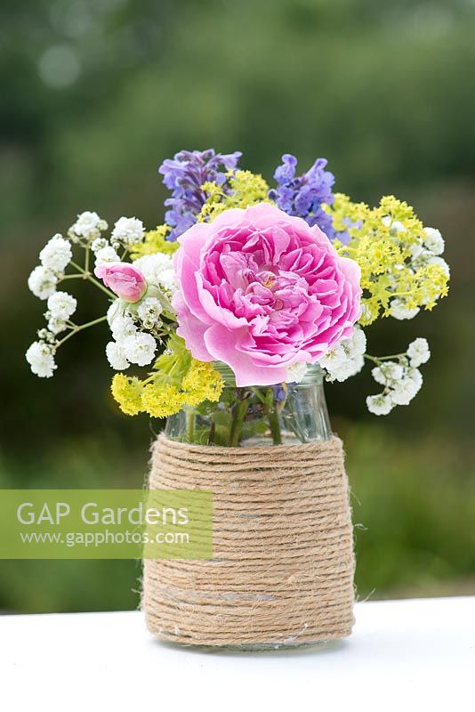 A colourful summer posie with pink rose, alchemilla, baby's breath and catmint in a glass jar decorated with twine.