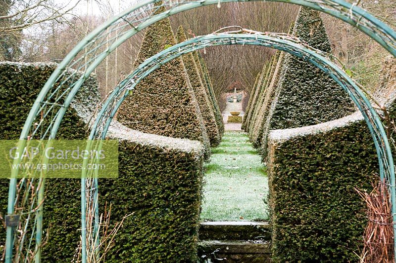 Rose arches frame a view along the garden's central axis flanked by clipped yew pyramids leading toward an obelisk at the far boundary