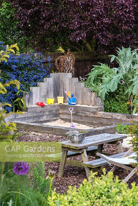 A sand pit and children's play area made from railway sleepers with floor covered with bark chippings. Behind, grey leaved cardoon, red leaved prunus and blue ceanothus.