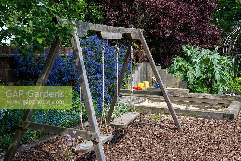 A wooden swing and sand pit made from railway sleepers in a children's play area with floor covered with bark chippings. Behind, grey leaved cardoon and blue ceanothus.