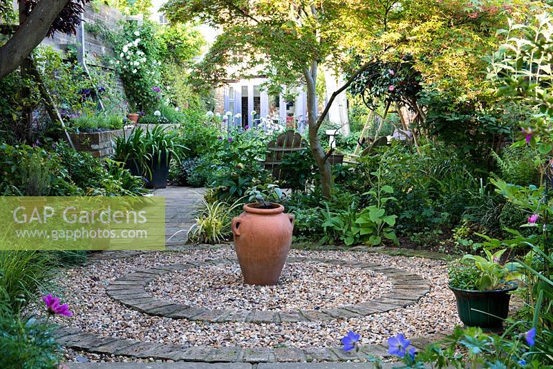 Circular courtyard made from circle of brick filled in with gravel, a terracotta urn at its centre. Surrounding borders planted with shade-loving hellebores, hardy geranium, dicentra, ferns, lamium and foxgloves. Behind on right, Japanese maple overhangs seat. 