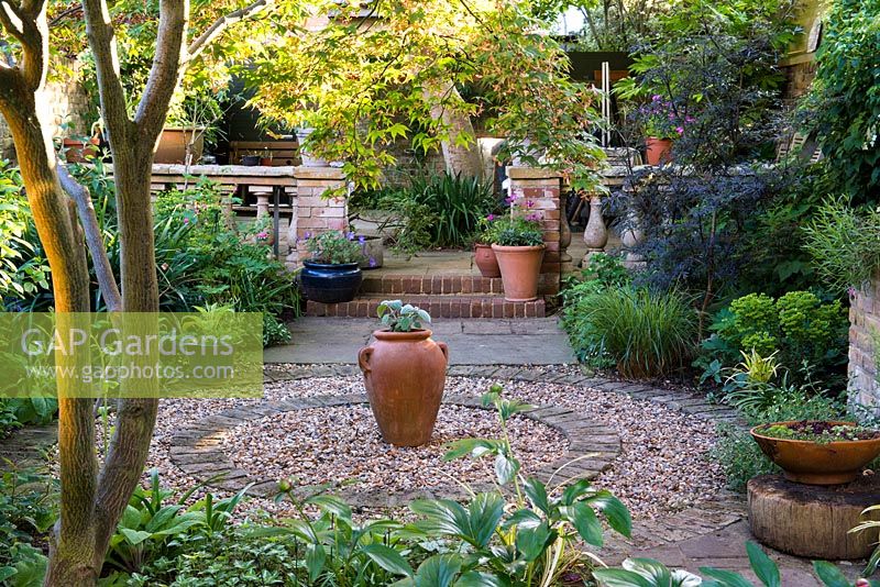 Circular courtyard made from circle of brick filled in with gravel, a terracotta urn at its centre. On left, trunk of Japanese maple. Borders planted with shade-loving hellebores, lamium, euphorbia, black elder, dicentra and foxgloves.