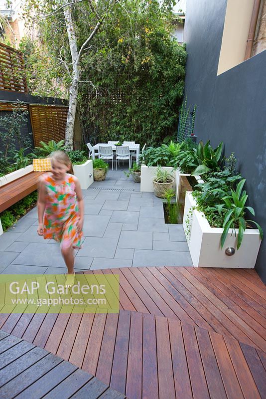 Young girl walks to timber decking in inner city courtyard with small dining area and back fence covered with Phylostachys nigra, black bamboo. Concrete planters with various shade loving plants seen. 