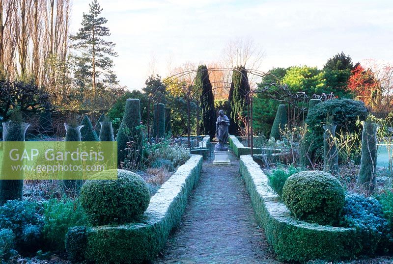 Walled garden with perennial parterre in winter frost. Central classical female figure Bacchus sculpture and circular metal framework for climbing vines. 