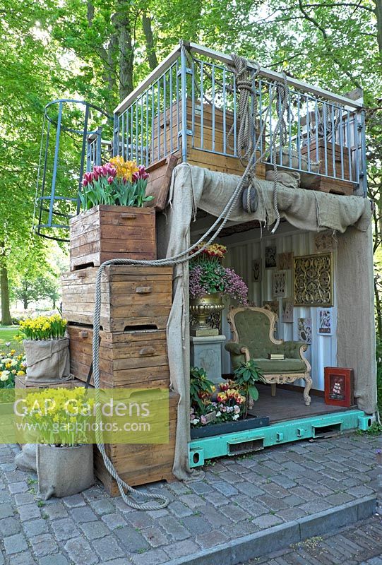 Tiny house in container with classic old aged furniture and pictures. Wooden boxes filled with Narcissus and Tulips. Terrace on top. Inspiration garden: Golden Age.