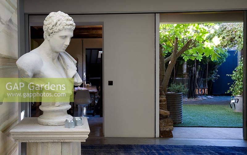 Interior with classical plaster bust and a view out to a courtyard garden with plants in containers. Full height glass doors open. 