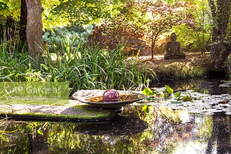 Pond with water lilies, buddha statue and clay bowl on the water's edge surrounded by waterside planting - July, Les Jardins de la Poterie Hillen