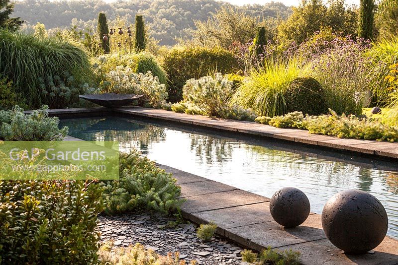 Modern pool edged with stone slabs and surrounded by mediterranean style planting - July, Les Jardins de la Poterie Hillen