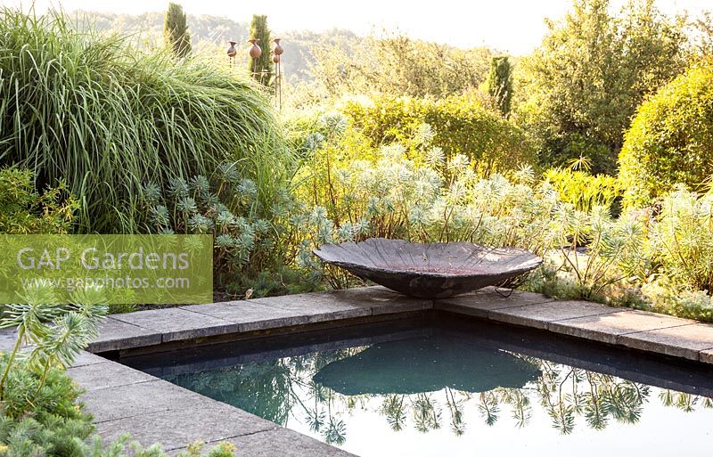 Modern pool edged with stone slabs and stone bowl surrounded by mediterranean style planting including Euphorbia characias, conifers and grasses - July, Les Jardins de la Poterie Hillen