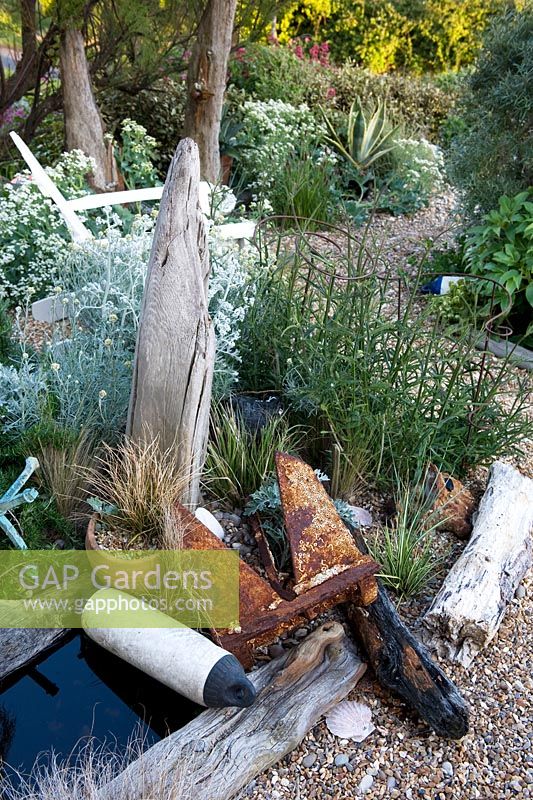 Seaside themed garden detail with white chair, boat anchor, buoys a small pool edged with driftwood, ornaments, Crambe maritima, verbena, agave and Gravel