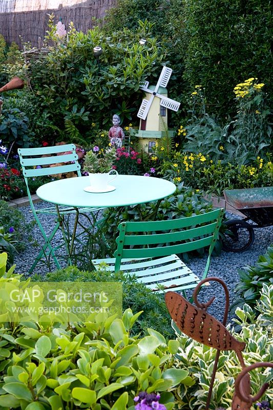 Garden packed with plants, ornaments and found objects including a windmill, boy figure, metal fish, wheelbarrow. Slate chip path. Cafe table. Colourful planting. 