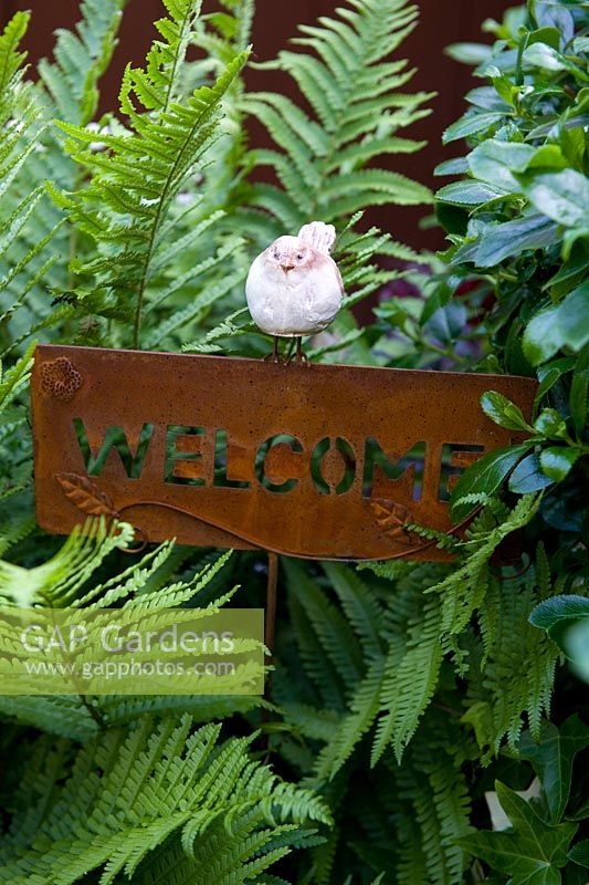 Little bird ornament perched on rusted steel welcome sign surrounded by fern foliage. 
