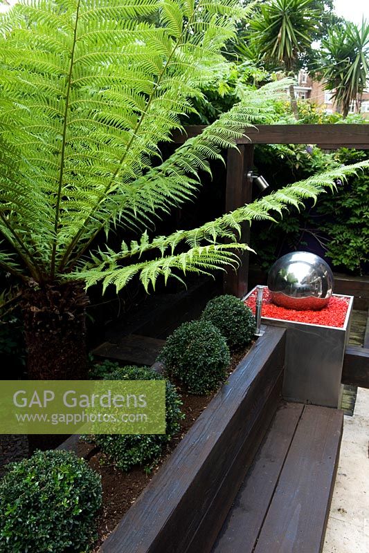 View from above of rail sleeper furniture and loggia with silver ball water feature, box balls in raised bed and a tree fern. 
