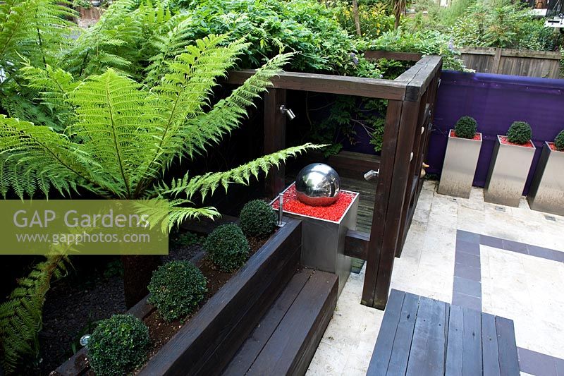 View from above of terrace, wooden furniture and loggia with silver ball water feature, box balls in aluminium containers. In raised bed a tree fern - Dicksonia antarctica.