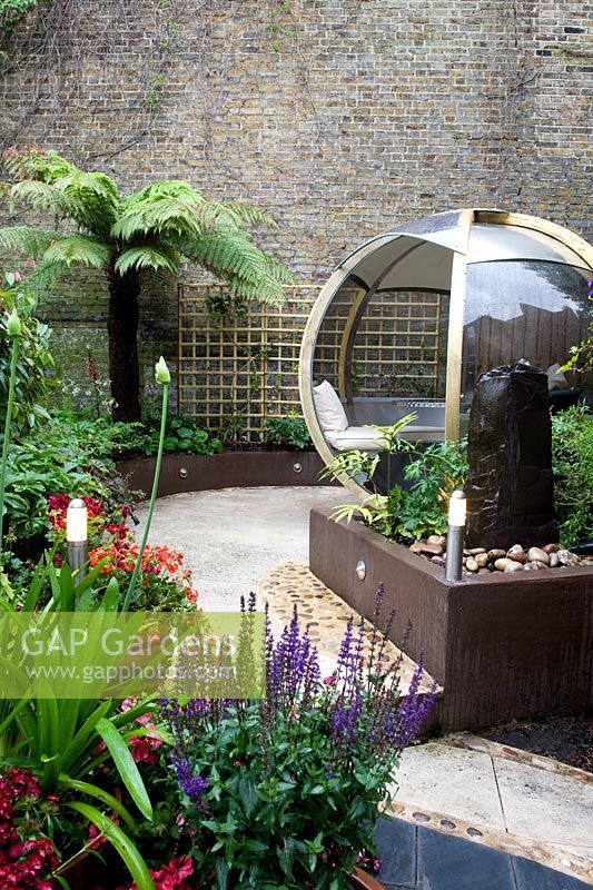 View of garden with brown painted low retaining walls and raised beds with colourful planting including salvia and agapanthus with a tree fern, high neighbouring wall, change of levels, hard landscaping, lighting posts and a large round wooden and perspex pod with seating and a table. Outdoor room. 