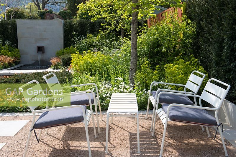 Seating area with white table and chairs under a Corylus colurna, Euphorbia robbiae, Anthriscus sylvestris and Anemone sylvestris, Graduate Gardeners: The Sunken Retreat Garden, RHS Malvern Spring Festival 2016