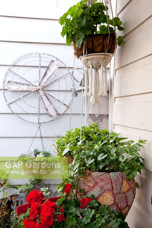 Detail of a front verandah showing a collection of pots and hanging baskets with a old fan blade and cover as a wall decoration, a retro cement pot planted with Spotted Dead Nettle, Lamium maculatum and a red flowered geranium - Pelargonium.